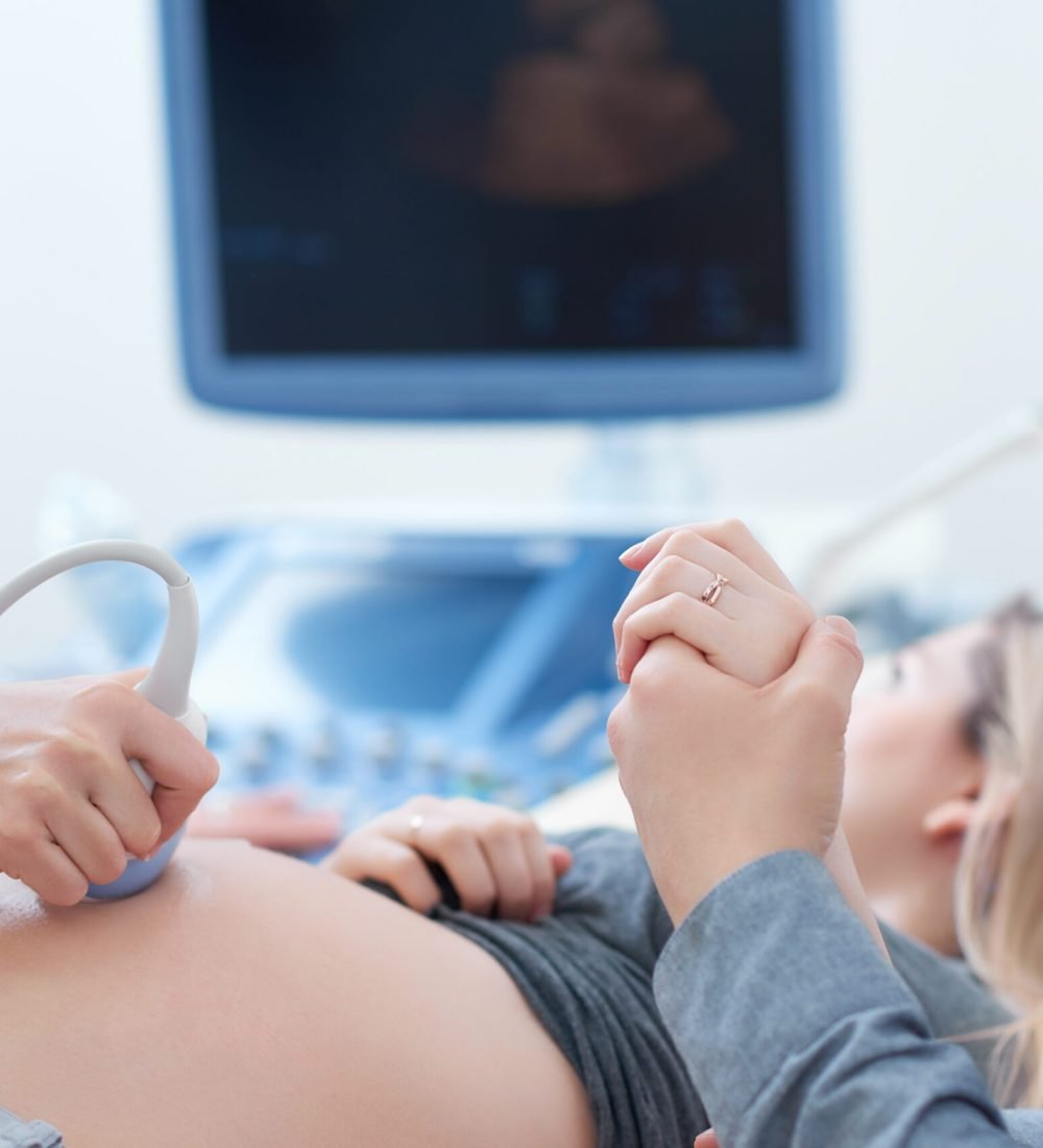 Female doctor touching stomach by tool of woman who lying,showing baby at screen of computer. Professional doctor using ultrasound equipment screening of pregnant woman. Husband holding hand his wife.