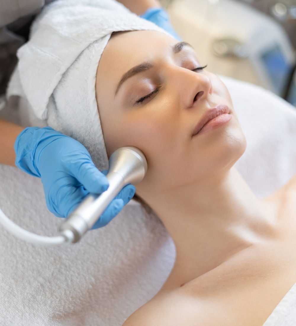 Young woman lying on cosmetologist's table during rejuvenation procedure. Cosmetologist take care about neck and face skin youthfull and wellness. Hardware face cleaning procedure
