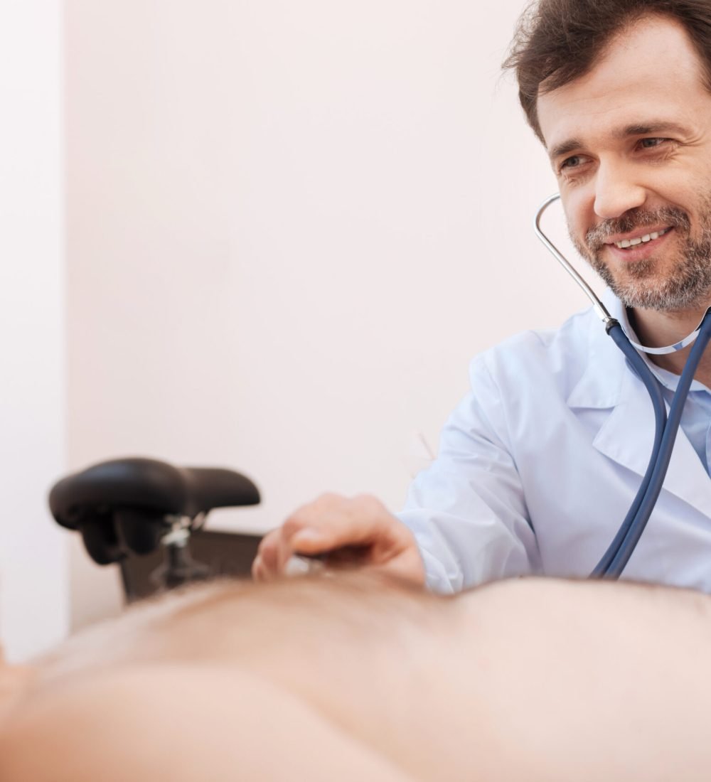 I will take care of you. Handsome nice competent doctor using a stethoscope hearing a mans heart while he lying on special chair